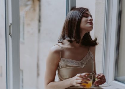Woman in a spaghetti strap dress savoring orange juice on a window sill, indoors.