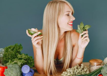 A smiling woman holding avocado and basil, surrounded by fresh vegetables and fruits for healthy eating.