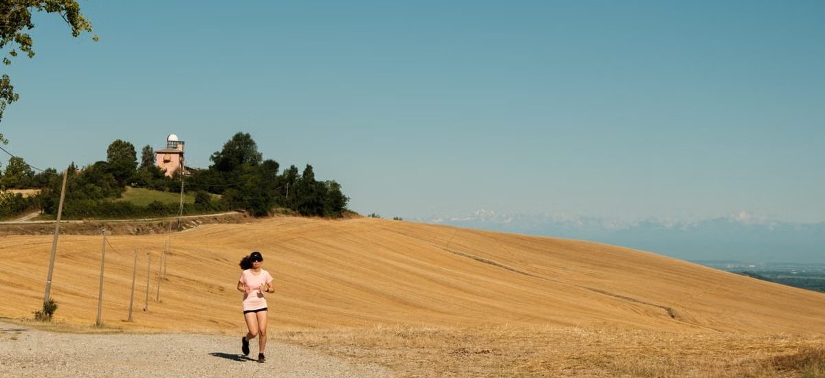 A woman runs on a gravel path through a sunlit rural landscape with dry fields and blue sky.