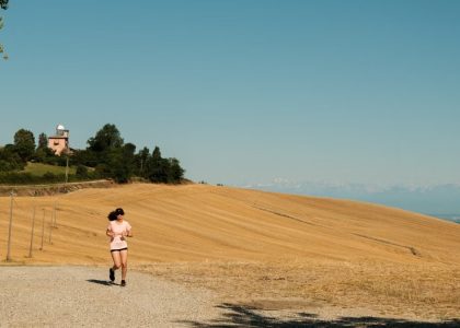 A woman runs on a gravel path through a sunlit rural landscape with dry fields and blue sky.